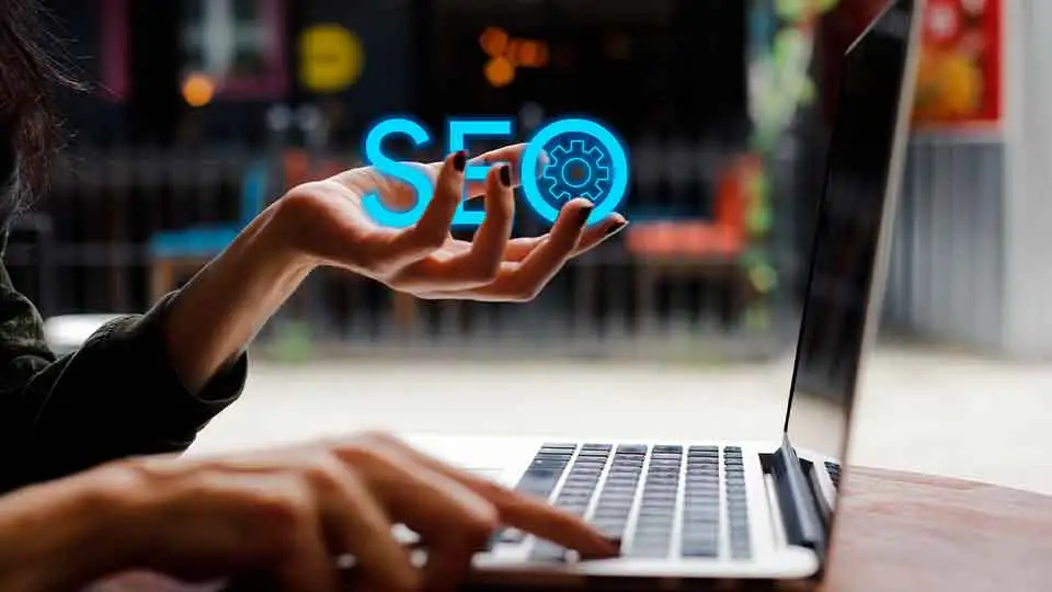 Bir kişi elini kaldırarak dizüstü bilgisayarında bir şeyler yazıyor, SEO harfleri ve dişli simgesi dijital olarak üst üste bindirilmiş ve bir b2b dijital pazarlama ajansı tarafından sunulan arama motoru optimizasyonu hizmetlerini simgeliyor.